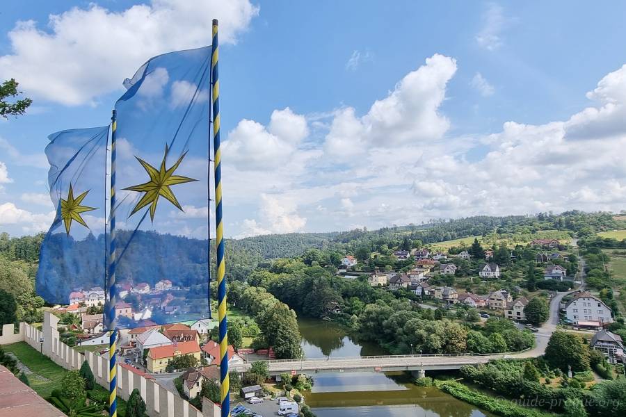 Two blue flags with yellow stars fluttering in the wind, positioned on a hilltop overlooking a small riverside town. The town lies along the Sázava River, surrounded by green hills and trees under a bright, partly cloudy sky.