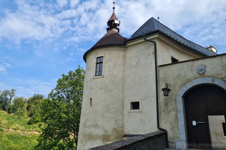 A section of a castle featuring a tall, round tower with a conical roof, set against a backdrop of greenery and a bright, partly cloudy sky. Trees and rolling hills can be seen in the distance.