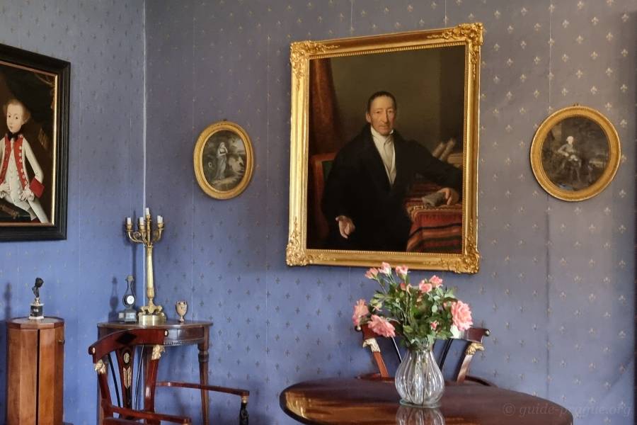 A photograph of a historical room in Český Šternberk Castle featuring a large portrait of Kaspar Maria Sternberg in an ornate frame. The portrait hangs on a wall with blue patterned wallpaper. Surrounding the central painting are smaller oval portraits. A round wooden table with a vase of pink flowers is positioned below the portrait.