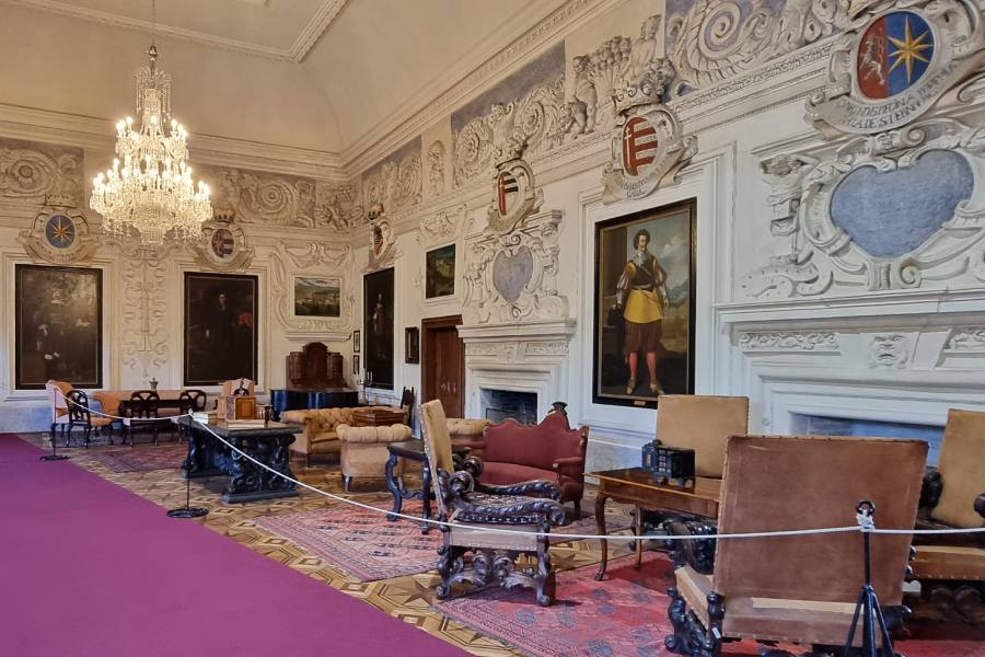 A grand Knight Hall inside a castle, featuring ornate white stucco walls adorned with intricate carvings and coat of arms. Large portraits hang between the decorations, while a sparkling chandelier illuminates the room. Antique furniture, including armchairs and tables, is arranged on a patterned rug over a polished floor, giving the space an opulent atmosphere.