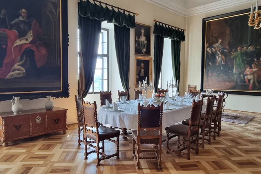 An ornate dining room inside a castle, featuring a long wooden table set with crystal candelabras and fine china. Large paintings hang on the walls, and tall, dark green curtains frame the windows, with sunlight illuminating the polished wooden floor.