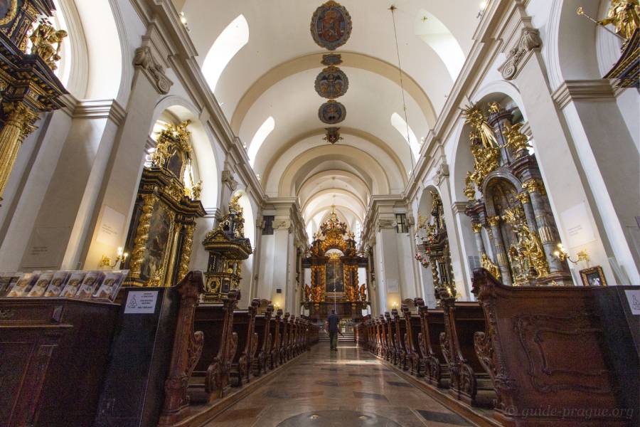 Interior of the Church of Our Lady Victorious in Prague, showcasing the grand baroque-style nave with wooden pews, ornate altars, and gilded decorative elements.