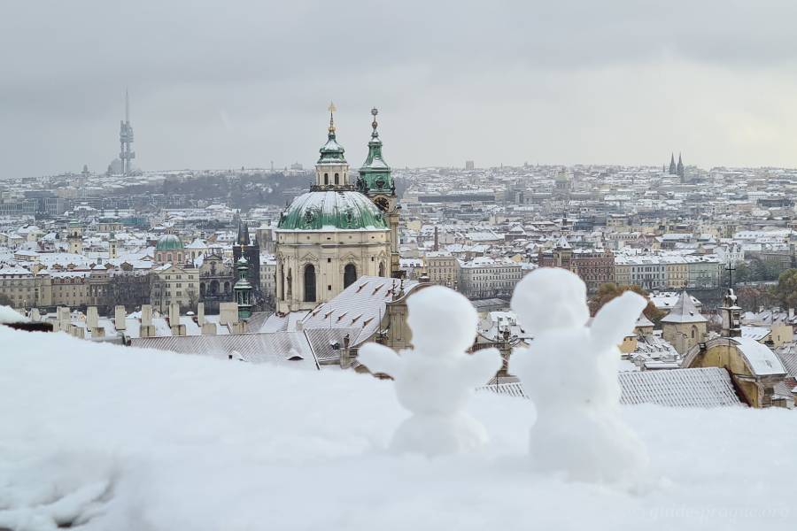 Two small snowmen overlooking Prague’s iconic skyline with St. Nicholas Church in the foreground and a wintery landscape in the background.