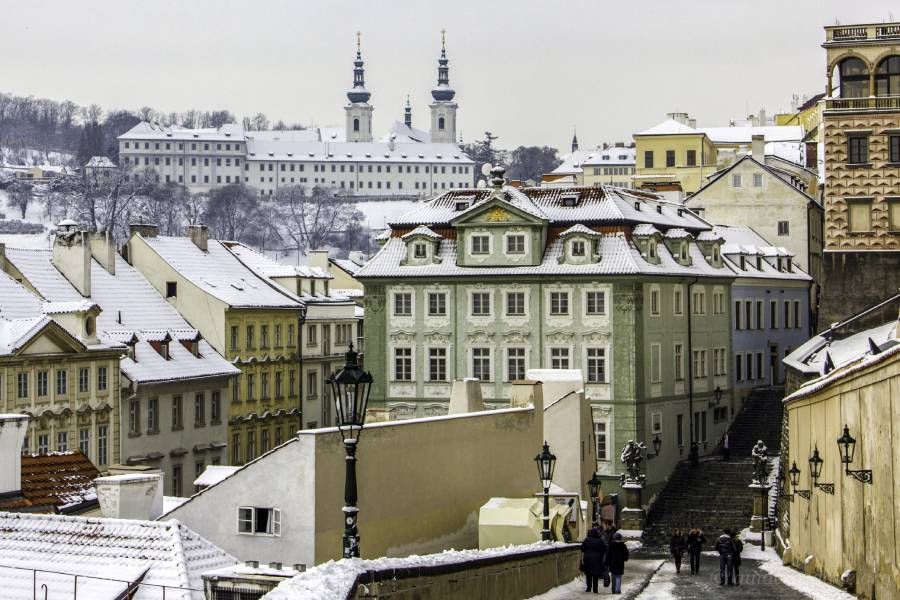 A panoramic view of Prague’s rooftops blanketed in snow, with St. Nicholas Church in the foreground and the iconic Žižkov TV Tower rising in the distance under a cloudy winter sky.