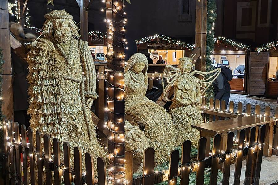 A beautifully crafted nativity scene made entirely of straw, featuring figures of Mary, Joseph, and an angel, surrounded by soft festive lights in a Christmas market setting in Prague.