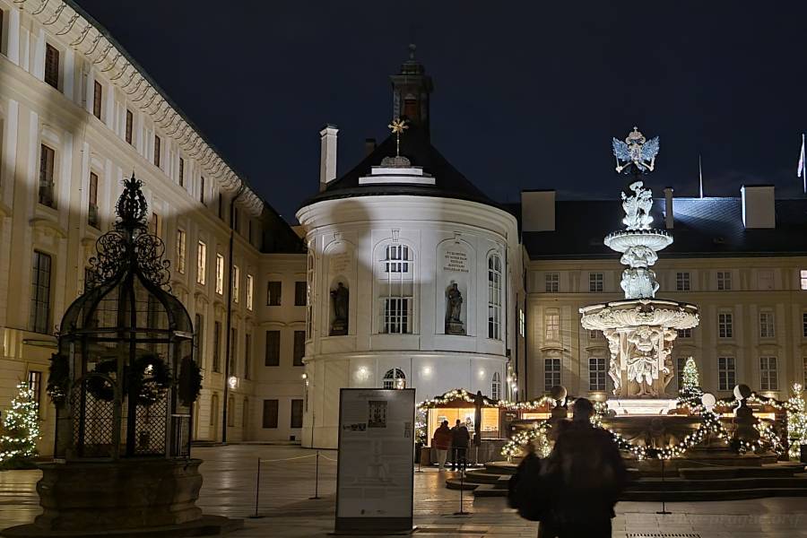 A beautifully illuminated courtyard at Prague Castle during Christmas, featuring festive lights, a decorated fountain, and historical architecture glowing against the night sky.