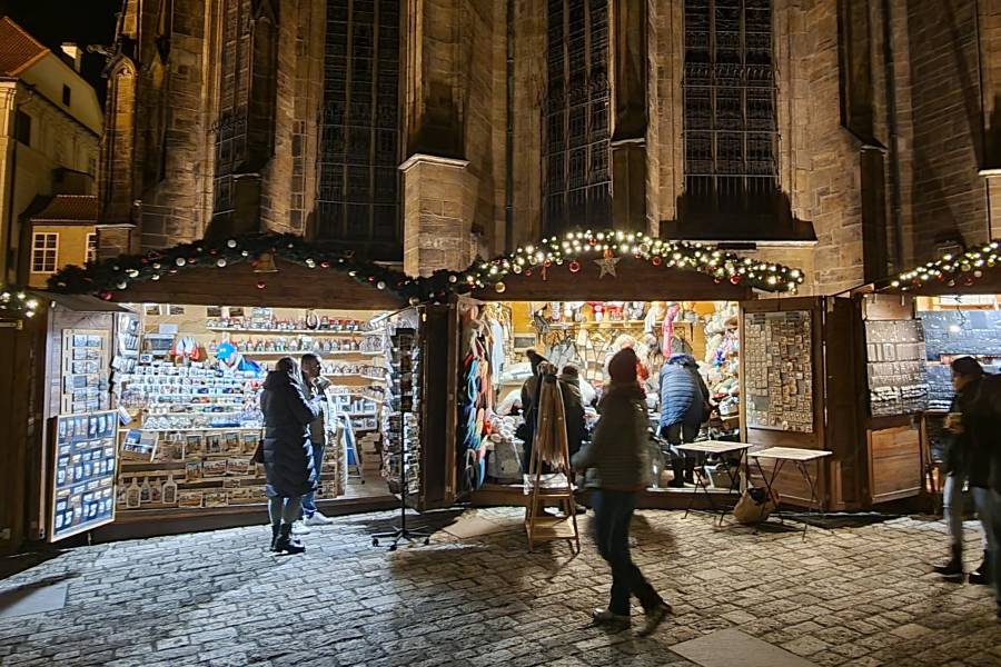 A cozy Christmas market stall in Prague, decorated with festive lights, offering holiday souvenirs and gifts under the soft glow of nighttime, with visitors browsing and enjoying the seasonal atmosphere.