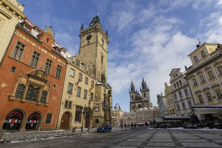 A bright winter morning at Prague’s Old Town Square, showcasing the iconic Astronomical Clock, colorful historic buildings, and the twin spires of Tyn Church, with a light dusting of snow covering the ground.