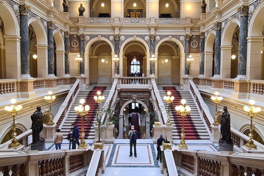 The majestic interior of Prague’s National Museum, featuring grand staircases, marble columns, ornate arches, and golden chandeliers, with visitors admiring the opulent Neo-Renaissance architecture.