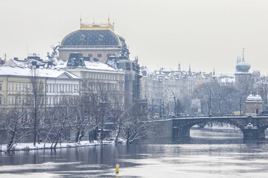 A scenic winter view of Prague's National Theatre with its golden rooftop, and the historic Lazansky Palace, home to Café Slavia, overlooking the snow-covered Vltava River.